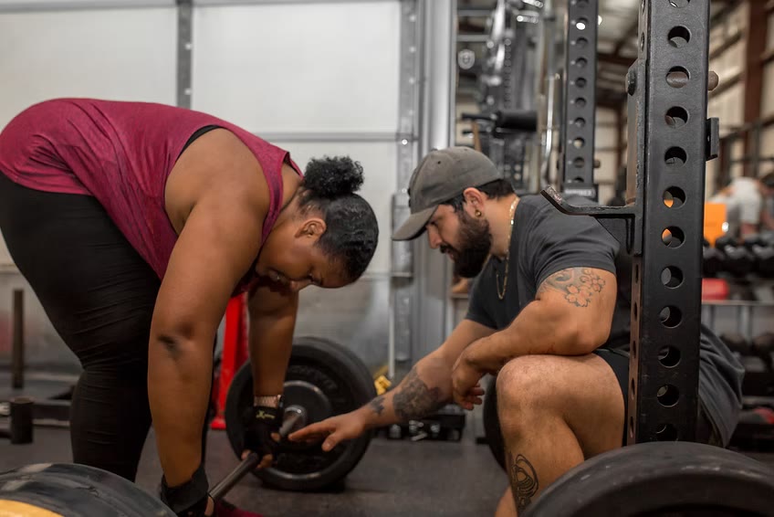 Martin kneeling beside a client to check her setup before a barbell lift