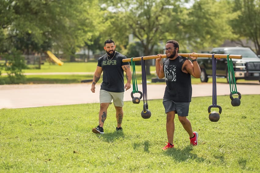 Two clients carrying a kettlebell-loaded bar across the grass outdoors